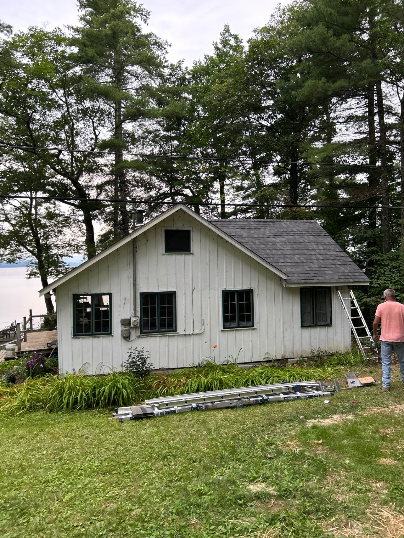 White lakeside cottage with old worn shingle roof, pines and water in background