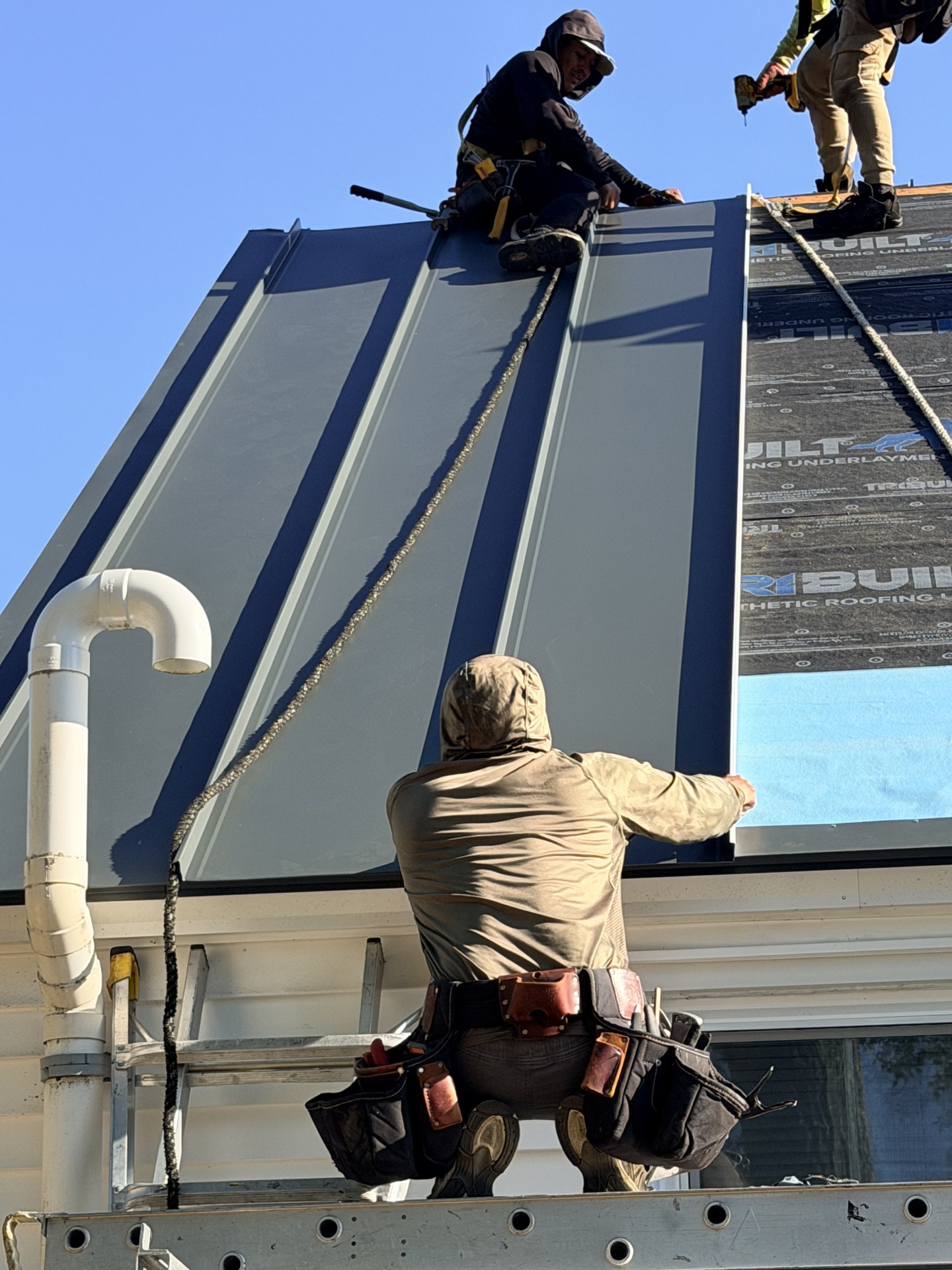 Roofers setting a charcoal Drexel standing-seam panel over synthetic underlayment