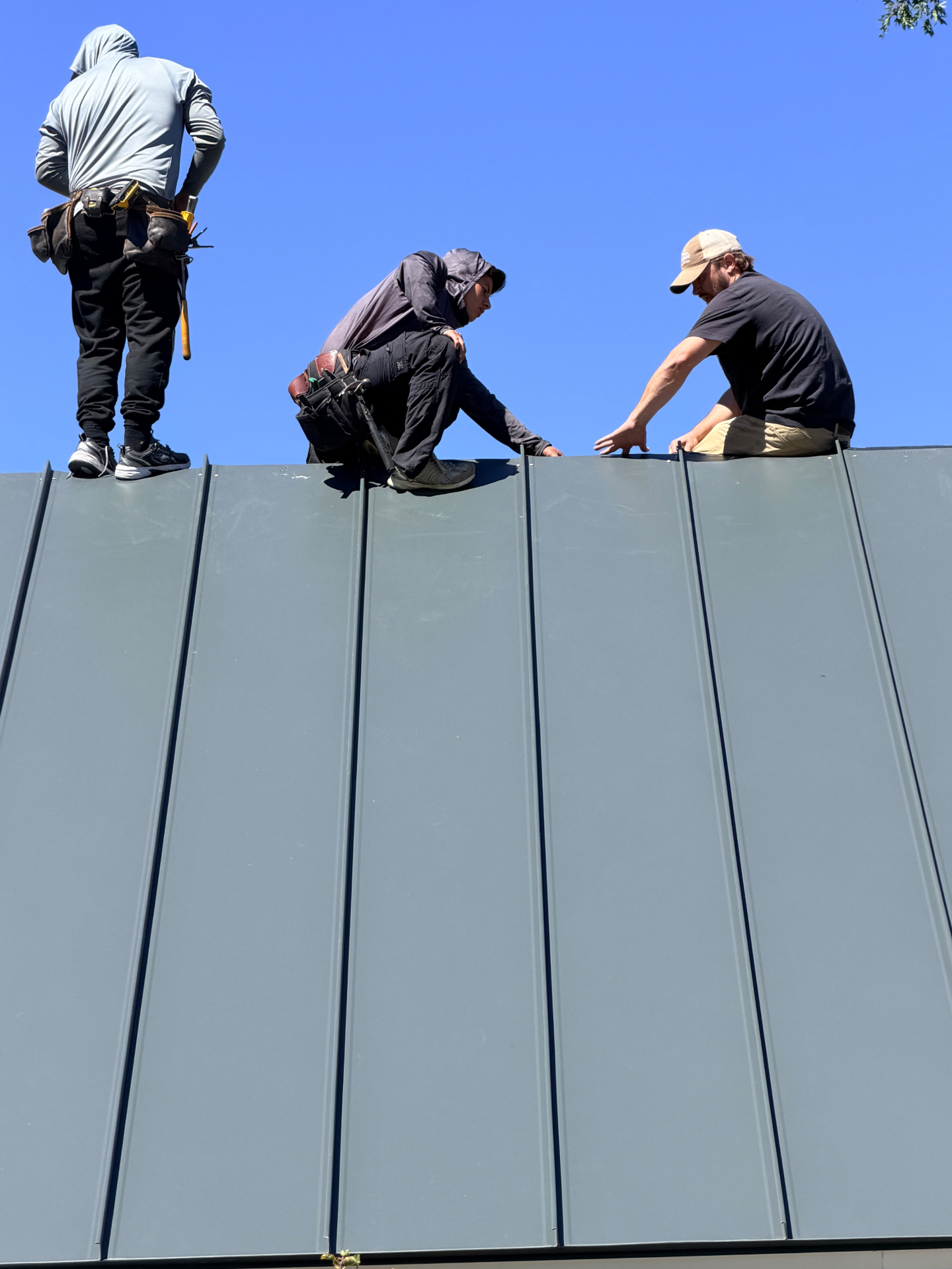 Three roofers locking standing seam panels into place on the charcoal metal roof