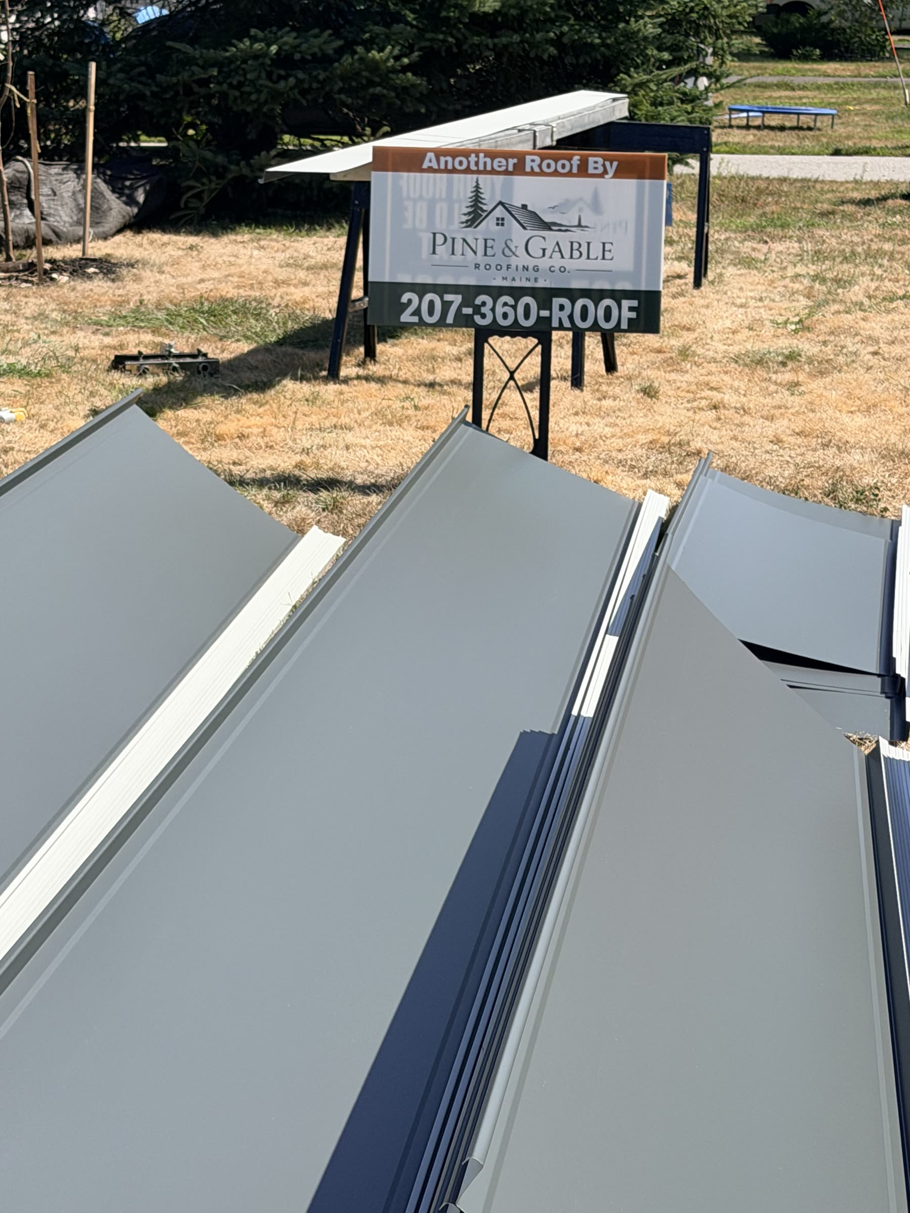 Long charcoal standing seam panels laid out on the lawn beside the Pine & Gable Roofing job sign
